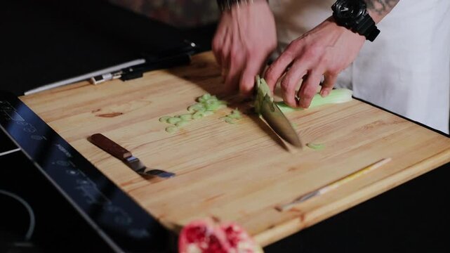Close-up over--shoulder view of cook's hands chopping celery on large light wood cutting board, chef's knife angled downward cuts pale-green stalk into thin rounds that scatter and form small pile