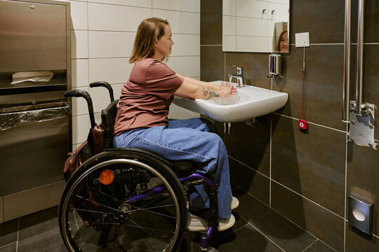 Young adult Caucasian woman with disability sitting in wheelchair washing hands at accessible sink in hotel bathroom. Image suitable for accessibility and healthcare campaigns