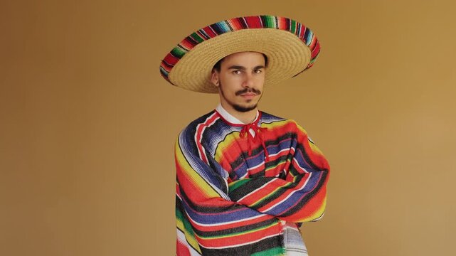 Man wearing wide-brimmed woven straw sombrero and vibrant striped serape lifts head, makes direct eye contact, and smiles while keeping arms crossed against smooth warm ochre studio backdrop, soft