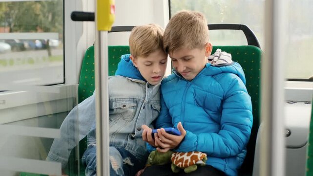 Two young boys sit side-by-side on green patterned bus or tram seat, leaning inward while older boy holds blue-cased smartphone and both look down at screen, with boys, children, sibling, smartphone