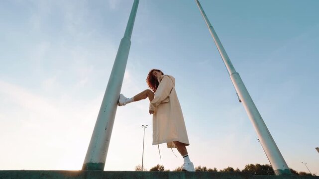 Young woman with curly red hair balances beside tall teal poles, one leg raised and one arm extended in confident fashion pose, with woman, balancing, pose, fashion detail, showing outdoor, lowangle