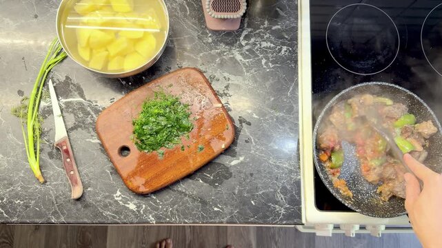 Overhead view of cook stirring meat and vegetable stew beside chopped parsley on cutting board.