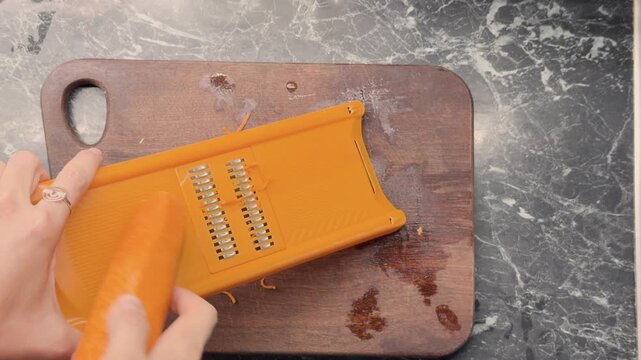 Hands grating fresh carrot into thin strips with julienne grater for Korean carrot salad.