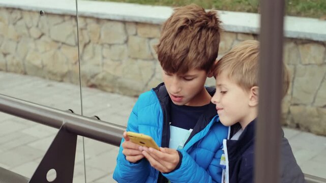 Two boys sit closely on outdoor bench while older boy holds yellow smartphone with both hands and younger boy leans in to view screen, visible action is mutual gaze shift toward device and