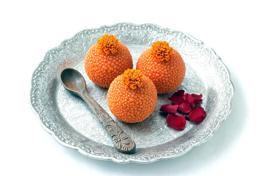 Three orange spherical sweets with marigold flowers on a decorative silver plate with a spoon and rose petals