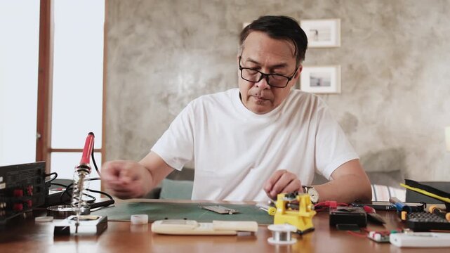 Mature Asian man concentrates on repairing home electrical appliance, disassembling and soldering circuit board, backache, bad posture, loosening up, taking a break, resting from his workshop hobby.