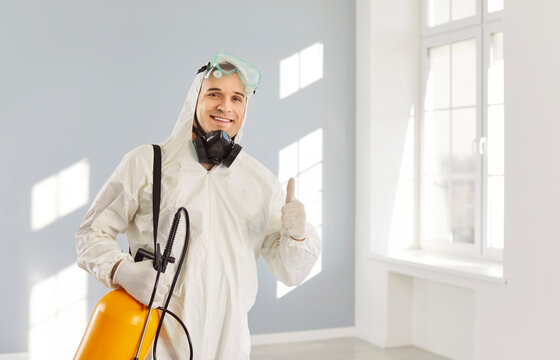 Exterminator giving thumbs up after insect extermination, sanitation work. Pest control service worker with sprayer, hazmat suit and mask smiling, standing in clean room after professional treatment