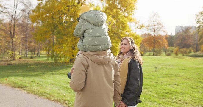 Family mother father daughter walking autumn park. Happy parents walk as a child rides on fathers shoulders near golden trees and green lawn. Theme shows family love, unity, and joy.