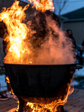 Large black cast iron cauldron boiling over an open fire with bright flames and smoke outdoors