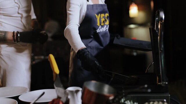 Female chef in foreground wearing glasses, white long-sleeve top and dark apron with yellow graphic extends gloved hands to place plated dish onto service surface while male chef in white jacket and