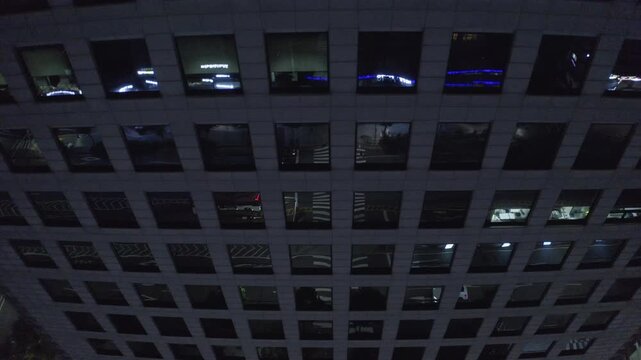 Geometric office tower windows mirror dim city streets as a drone ascends along the facade at dark.