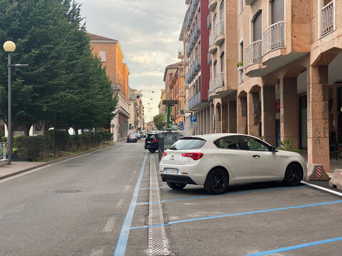A modern white Alfa Romeo Giulietta hatchback is parked on a city street with marked blue parking bays and trees lining the sidewalk