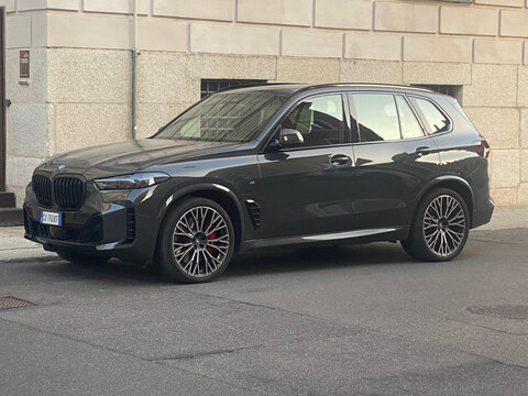 A sleek, dark gray BMW X5 M Competition SUV is parked on an asphalt road in front of a stone building