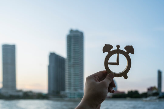 Real estate timing and property investment concept with hand holding wooden alarm clock against river and modern skyscraper buildings for best time to buy home, mortgage and urban living.