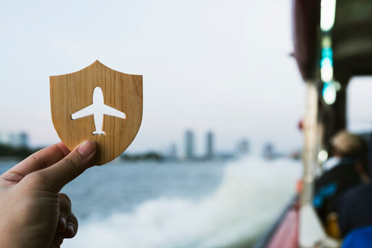 Travel insurance and holiday journey protection concept with hand holding wooden shield with airplane cutout on passenger boat over water for flight safety, vacation and transit coverage.