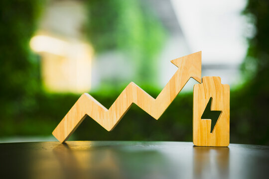 Energy cost increase and electricity tariff concept with wooden upward trend arrow pointing to battery block with lightning symbol on table against green bokeh for power expense growth.