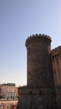 Stone tower of Castel Nuovo rises above the city of Naples under a clear daytime sky in a vertical composition.