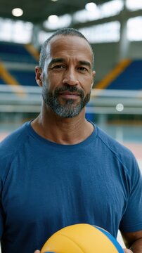 Male volleyball player with a beard holding a yellow and blue volleyball inside an indoor court with bleachers and a sports facility in the background