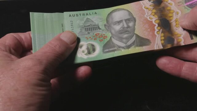 Mature hands bend and fan a thick bundle of Australian 100 notes against a dark surface, showing texture and detail.