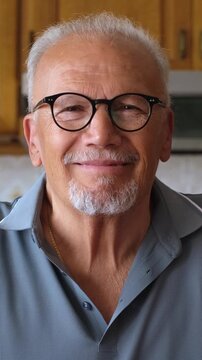 Close-up portrait of an elegant senior man with grey hair and a goatee, wearing glasses and a blue polo shirt, looking at the camera and starting to smile genuinely in his kitchen