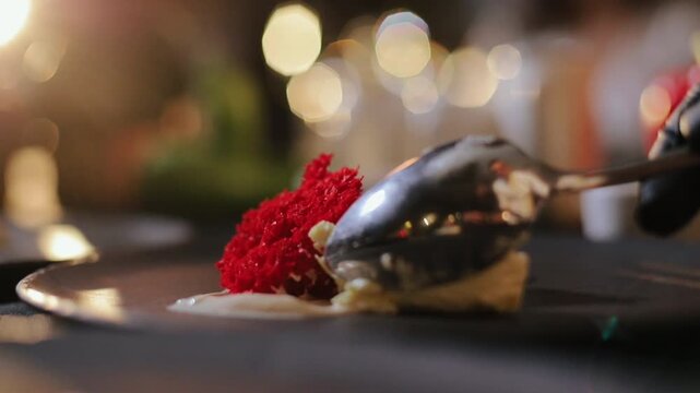 Metal spoon lifts and adjusts plated dessert consisting of moist chocolate roll with cream filling and bright red textured sponge resting on pale cream sauce on glossy dark plate, with dessert, spoon