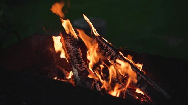 Flames dance around several logs in a fire pit as they burn brightly. The bright orange and yellow hues of the fire create a warm glow against a dark background