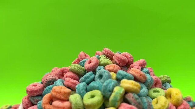 Colorful cereal rings fill a red bowl against a bright green background. Additional cereal pieces surround the bowl, creating a vivid closeup food composition.