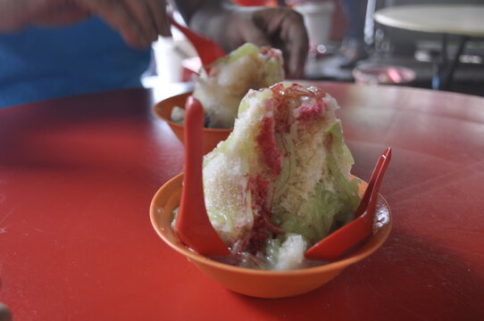 Traditional Malaysian shaved ice dessert (Ais Kacang and Cendol) with colorful syrups and green jelly, served in orange bowls on a red table at a local street food stall in Kuala Lumpur Chinatown. ABC