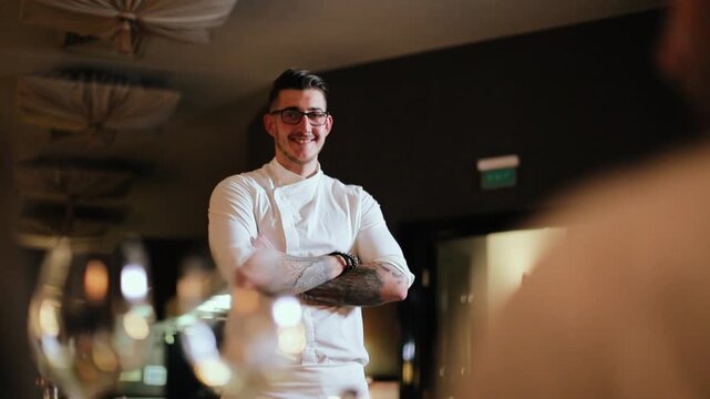 Waist-up view of male chef in white jacket standing inside dining room with arms crossed, shifting from neutral expression to friendly smile while remaining stationary, with chef, man, portrait
