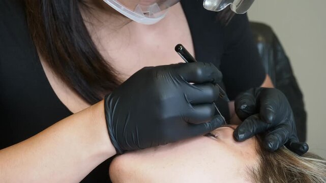 Cosmetic specialist in black nitrile gloves performs a precise eyebrow tattoo treatment on a reclining client, using a fine pigment application tool, permanent makeup procedure, extreme close-up.