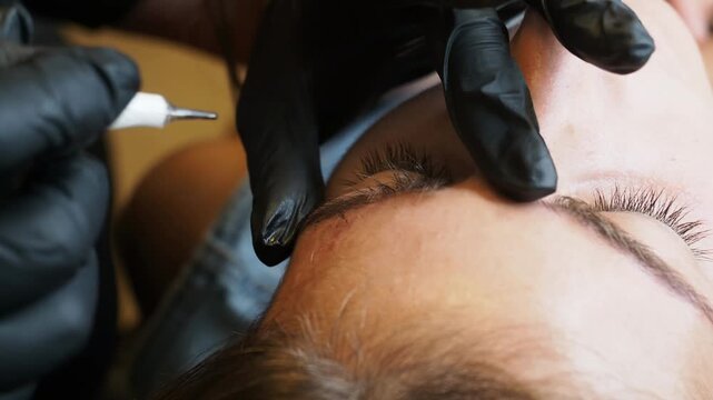 Eyebrow tattoo specialist in black nitrile gloves applies permanent pigment along the brow line with a precision needle during a PMU procedure, static extreme close-up.