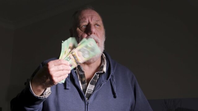 Elderly man in hoodie fans a thick stack of green currency under harsh side lighting, suggesting personal finance stress or windfall.