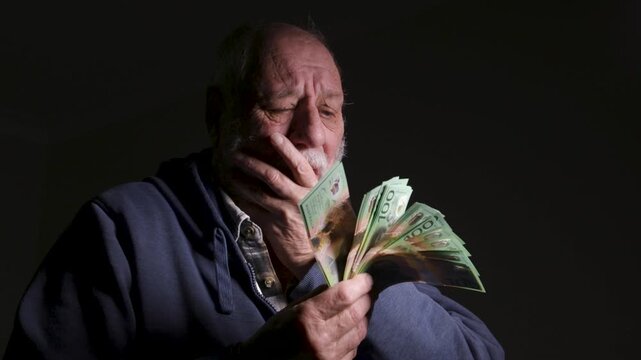 Anxious elderly man in hoodie counts a thick bundle of green banknotes, reacting with concern under dramatic side lighting.