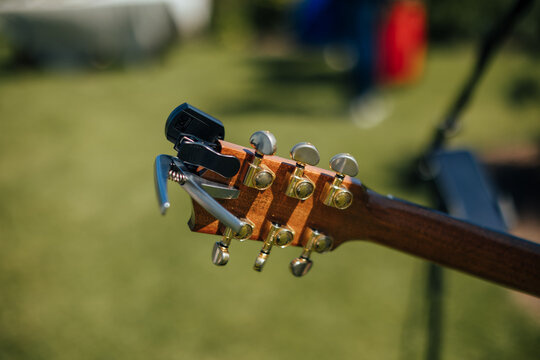 Close-up of a wooden acoustic guitar headstock with metal tuning pegs and a capo attached near the top, photographed outdoors against a softly blurred green background with copy space
