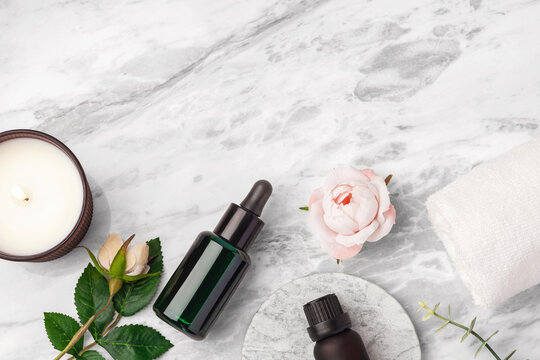 Green oil serum dropper bottle, essential oil bottle, burning candle and pink rose flowers on marble table background, top view