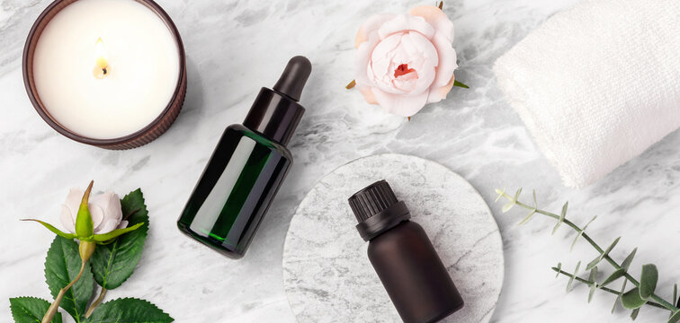 Top view of green oil serum dropper bottle, essential oil bottle, burning candle, white towel, and pink rose flowers on marble table, closeup