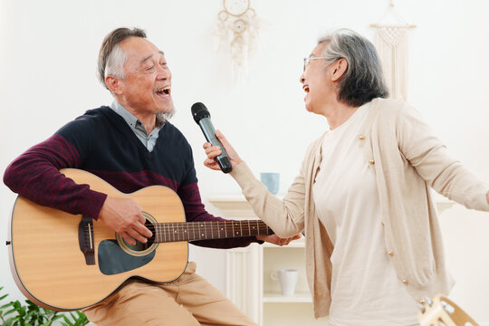 Cheerful, joyful senior Asian couple singing, dancing, playing guitar with joy, fun and happiness in modern, cozy living room at home. Active lifestyle, healthy retirement and mental wellbeing concept