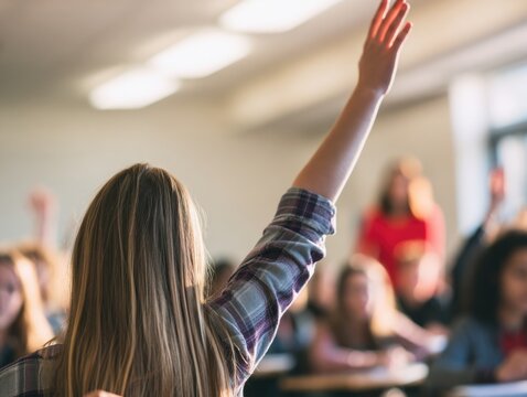 A student with long hair raises their hand in a classroom filled with attentive peers and a teacher in the background.