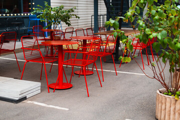 Obraz premium A row of red chairs surrounds several circular tables in an outdoor seating area. The space features potted plants and is set on a paved surface in a city environment