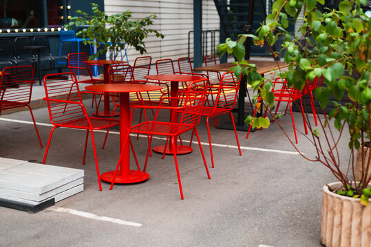 A row of red chairs surrounds several circular tables in an outdoor seating area. The space features potted plants and is set on a paved surface in a city environment
