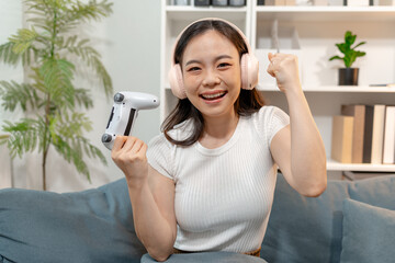 Young woman wearing wireless headphones while playing video games with a game controller at home. Excited gamer enjoying online gaming, entertainment, competition and modern digital lifestyle. © Imagestoyou