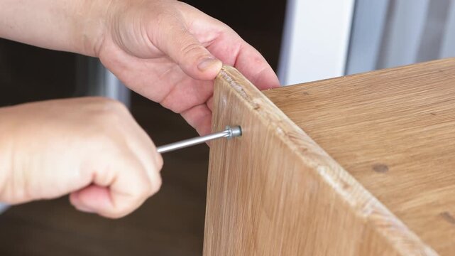 Close-up real-time shot of hands screwing a bolt into a wooden shelf with a hex wrench during DIY home furniture assembly. Ideal for flat-pack furniture tutorials, woodworking guides, home improvement