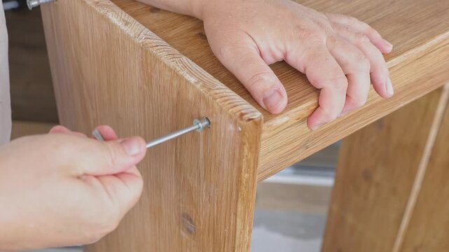 Close-up real-time shot of hands screwing a bolt into a wooden shelf with a hex wrench during DIY home furniture assembly. Ideal for flat-pack furniture tutorials, woodworking guides, home improvement