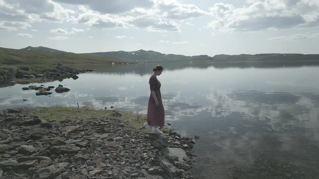 Aerial drone shot flying away and up from a young woman in a dress looking at camera on a mountain lake shore with sky reflection, flat log profile
