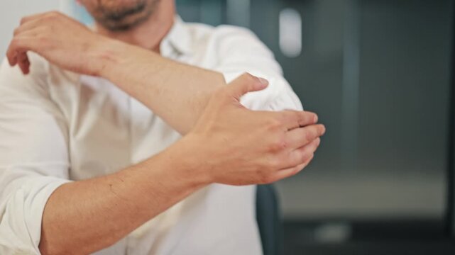Close-up video shot of a man in a white shirt experiencing acute pain in his elbow and forearm while working in an office. He holds the affected arm with his other hand, rubbing the joint.