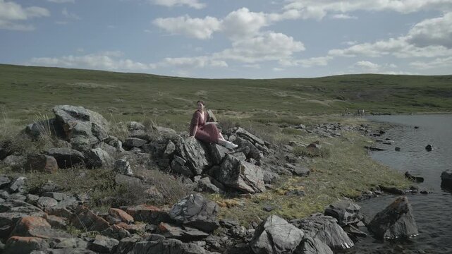 Low altitude aerial shot flying backward and away from a woman sitting on a lakeside rock to reveal a majestic mountain valley, flat log profile