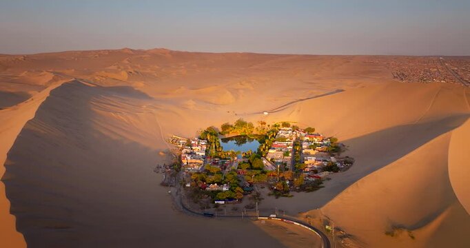 High aerial arc at sunrise of scenic Huacachina desert oasis amongst dunes, Peru