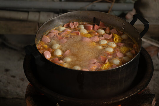Close-up of hot soup with sausages, corn and fish balls simmering in a large pot on charcoal stove