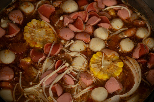 Top view of a rich flavorful hotpot with sliced sausages, fish balls, fresh enoki mushrooms and corn
