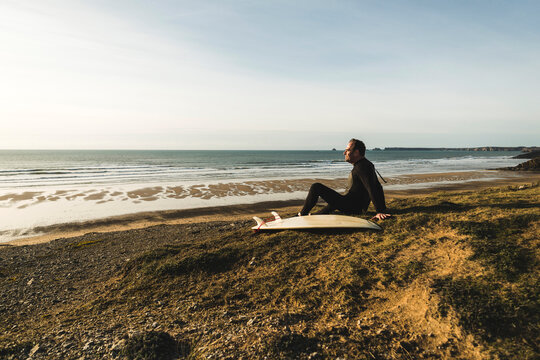 France, Bretagne, Finistere, Crozon peninsula, man sitting at the coast with surfboard